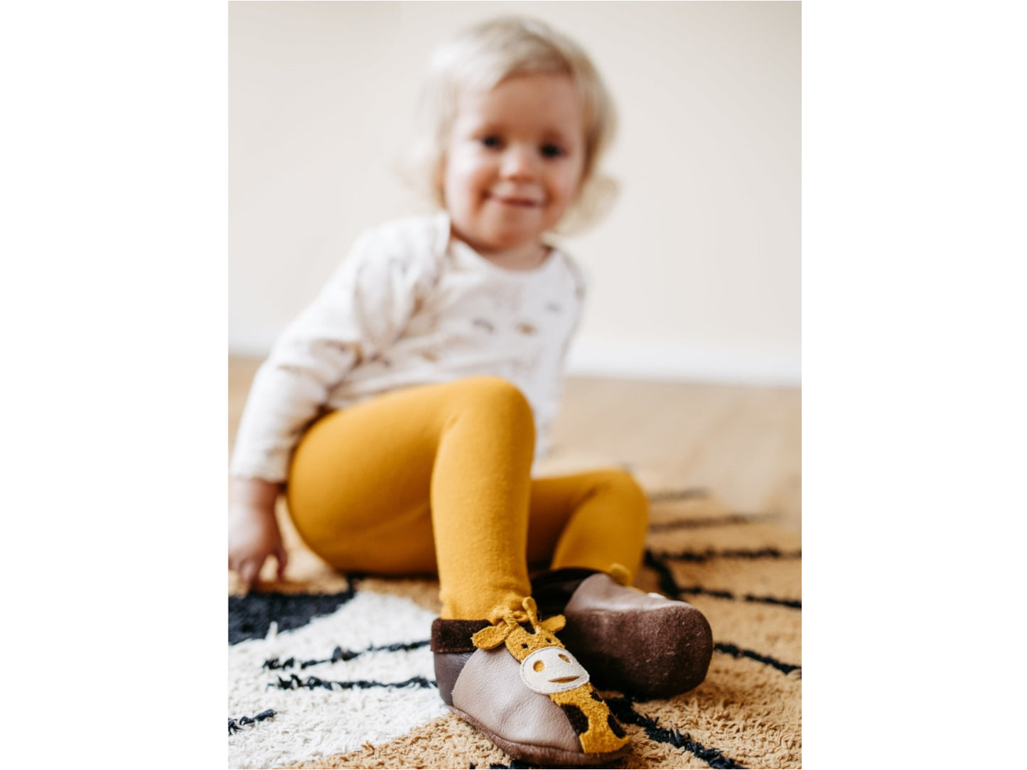 A smiling toddler sits on a cozy rug wearing mustard leggings and adorable giraffe-themed leather slippers.