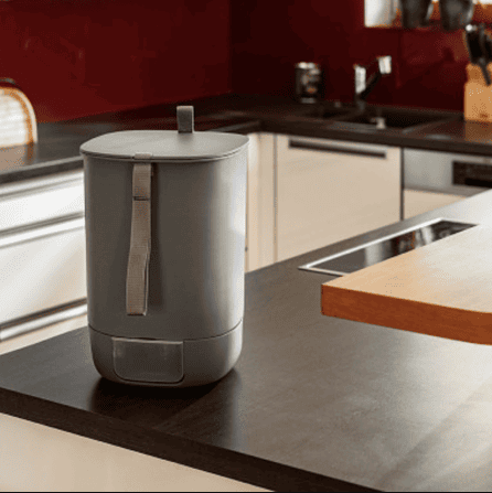 Grey Bokashi composting bin with handle placed on a modern kitchen countertop in a warmly lit interior.