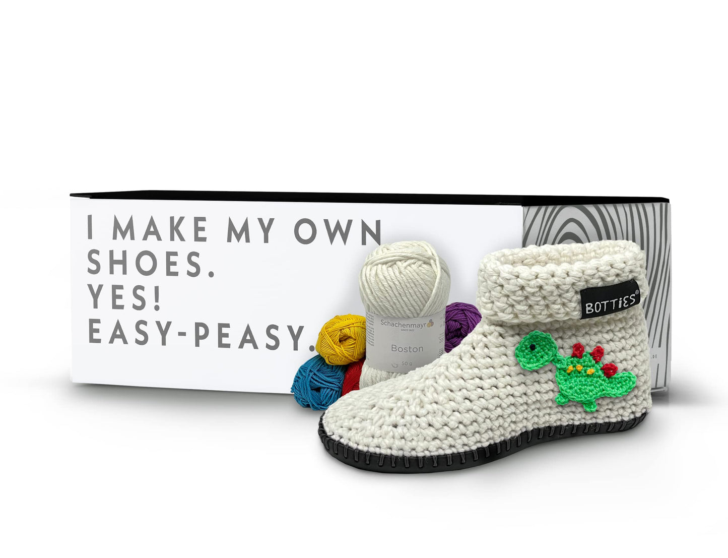 Botties crochet shoe DIY kit with a wool slipper, black sole, dinosaur applique, colorful yarn in front of a box