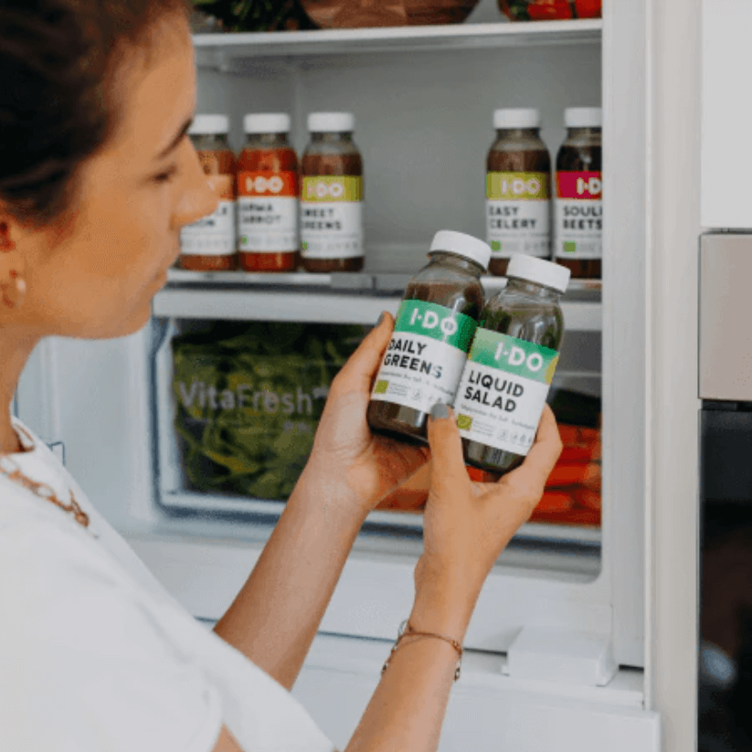 Woman holding I DO organic juice bottles, Daily Greens and Liquid Salad, in front of an open fridge filled with healthy juices.