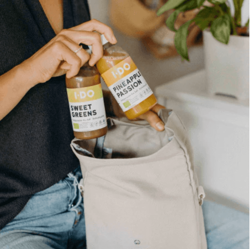 Person putting I DO organic juice bottles, Sweet Greens and Pineapple Passion, into a beige bag, promoting healthy lifestyle.