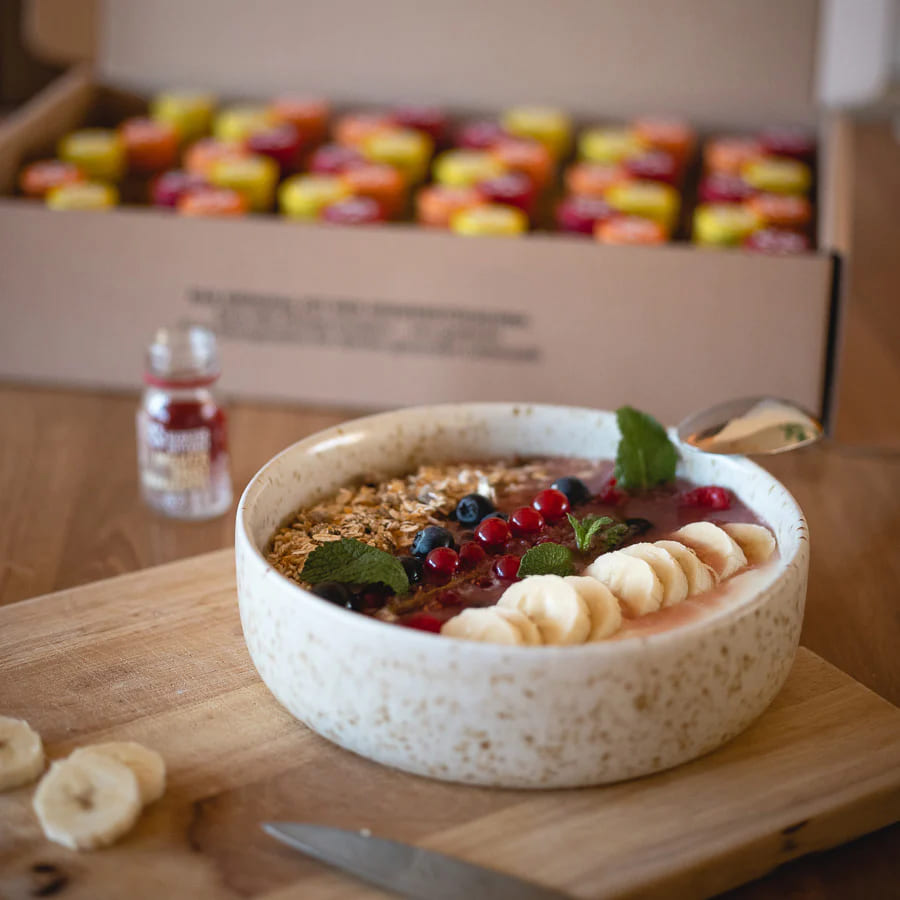 Smoothie bowl with fruit and granola next to a Kloster Kitchen ginger shot and a box of shots in the background.