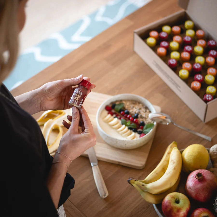 Smoothie bowl with fruit and granola next to a woman holding a Kloster Kitchen ginger shot