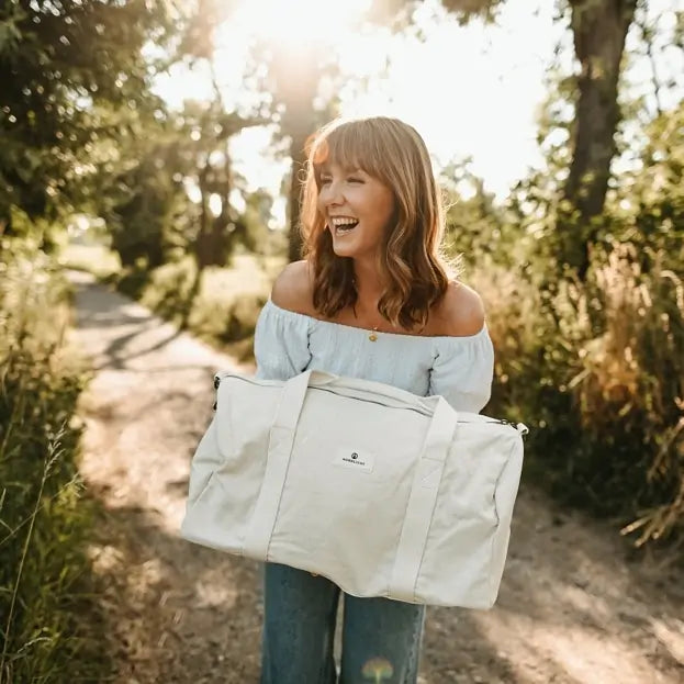 Smiling woman holding Nordlicht organic cotton bag on a sunny path, perfect for eco-friendly weekend adventures
