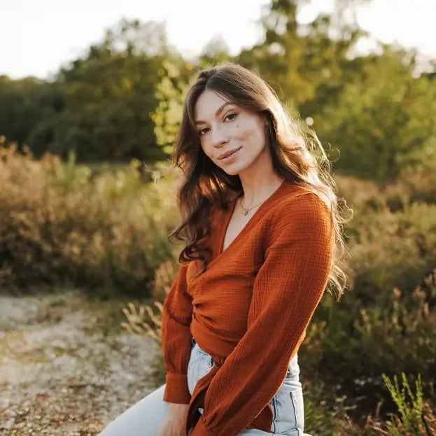 A woman in a rust-colored muslin wrap blouse poses outdoors in soft golden light casual, elegant, and naturally radiant.