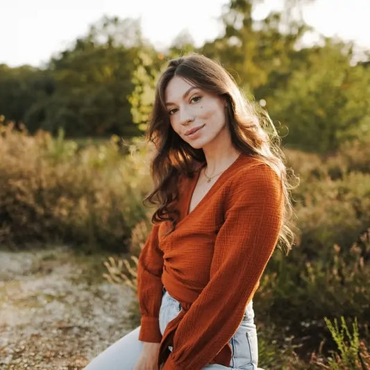 A woman in a rust-colored muslin wrap blouse poses outdoors in soft golden light casual, elegant, and naturally radiant.