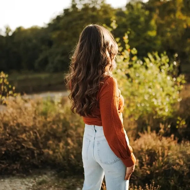 Woman in rust muslin blouse and jeans stands in golden field, surrounded by trees and soft autumn light.