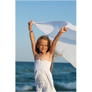 Girl on the beach joyfully waving a white organic cotton cloth, capturing summer freedom, play, and pure childhood bliss.