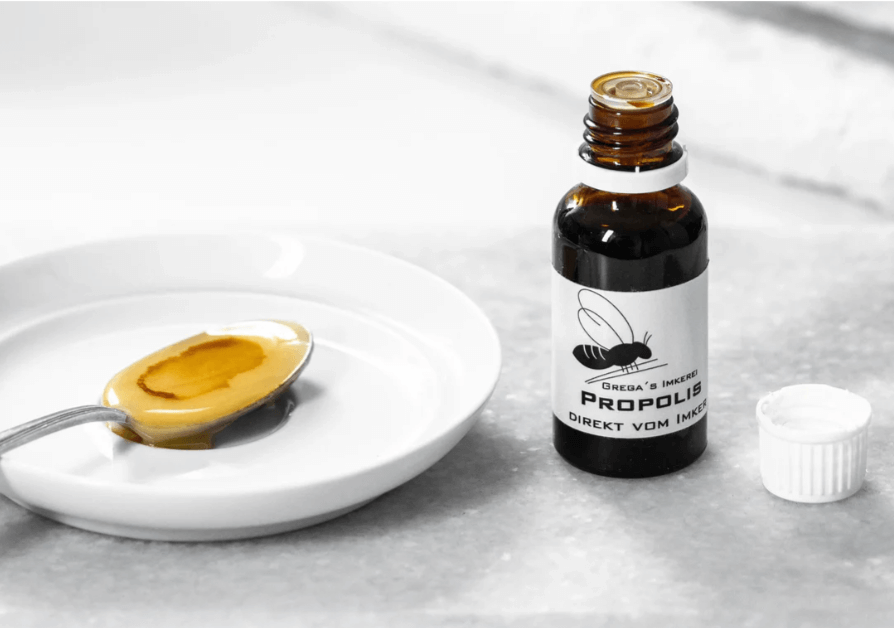 Handmade Propolis extract bottle by Grega's Imkerei, placed next to a spoon with honey on a white plate.