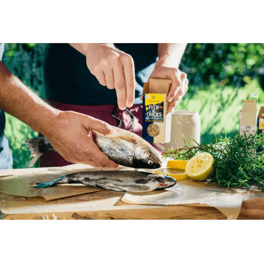 Hands seasoning fresh fish with Sonnentor Fish & Chicks grill seasoning, surrounded by lemon and herbs.