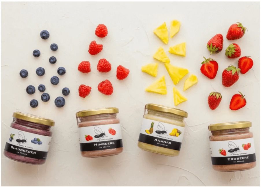 Four jars of honey fruit spreads with fresh blueberries, raspberries, pineapple, and strawberries on a white background.
