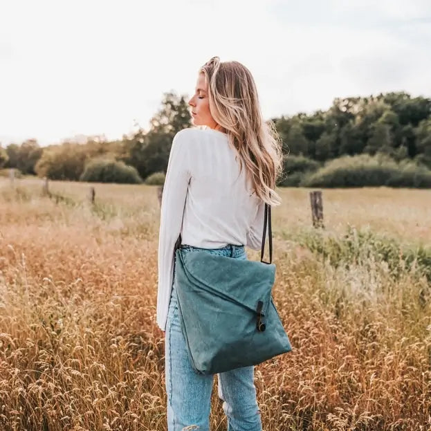 Woman outdoors holding a turquoise canvas handbag with leather strap, wearing jeans and white top.