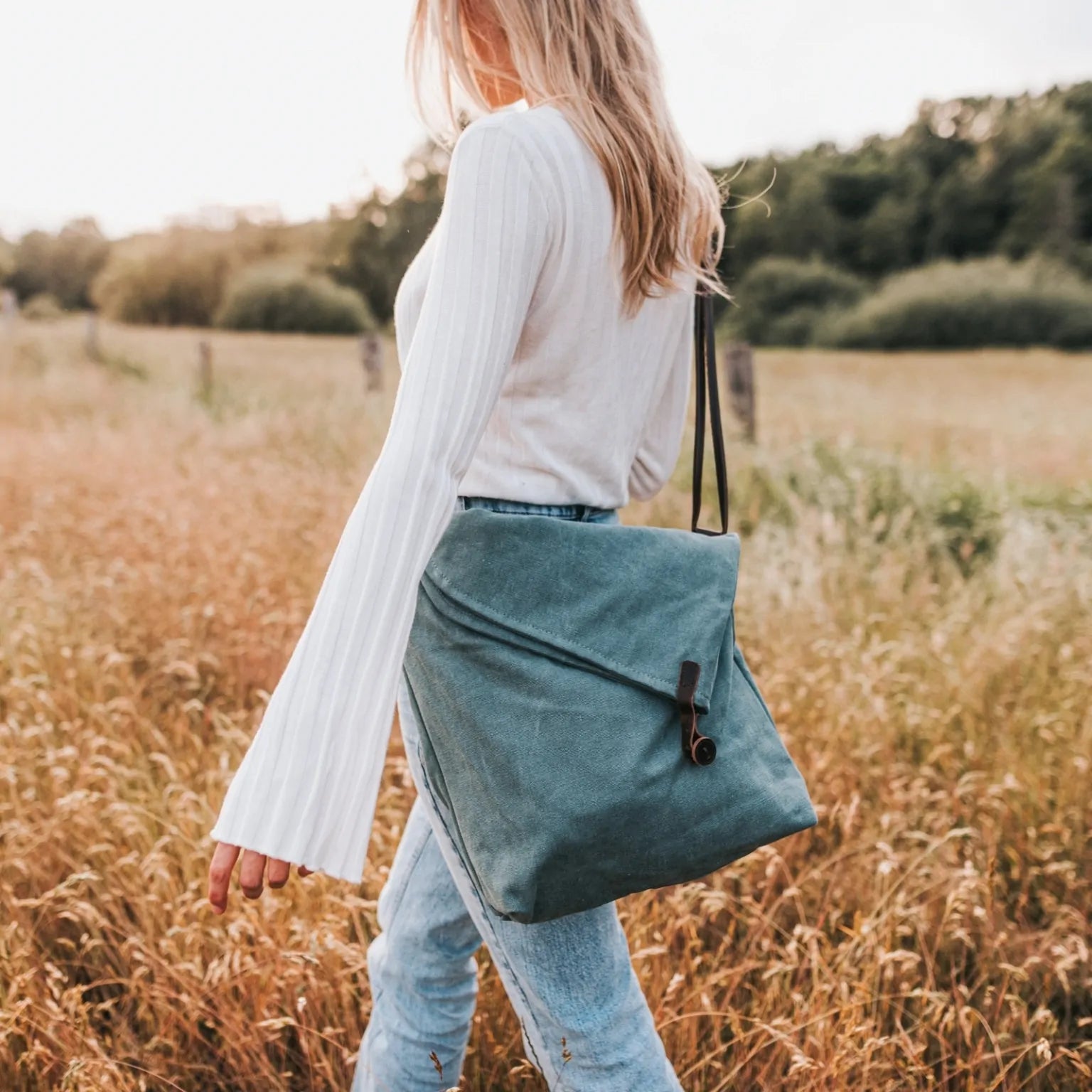 Woman outdoors holding a turquoise canvas handbag with leather strap, wearing jeans and white top.