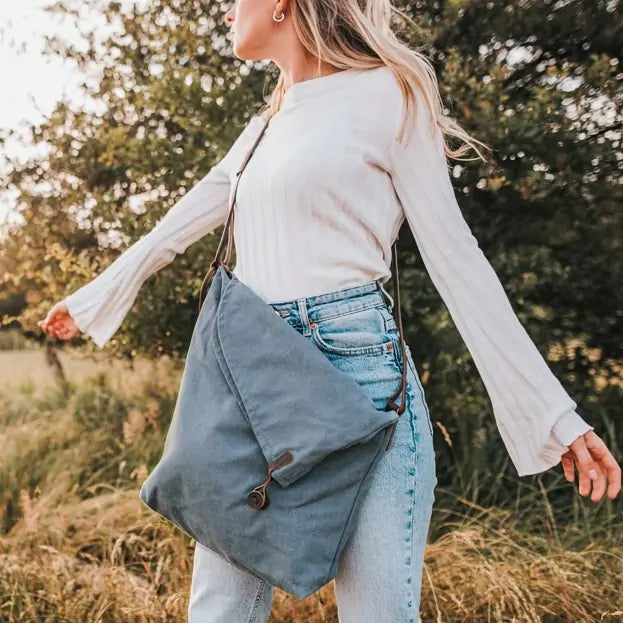 Woman outdoors holding a turquoise canvas handbag with leather strap, wearing jeans and white top.