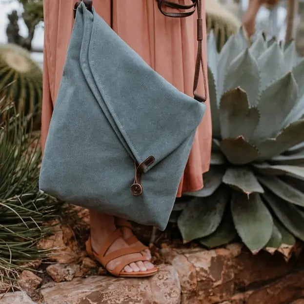 Turquoise canvas handbag with leather strap, worn crossbody by a woman in sandals and dress, outdoors near agave plants.