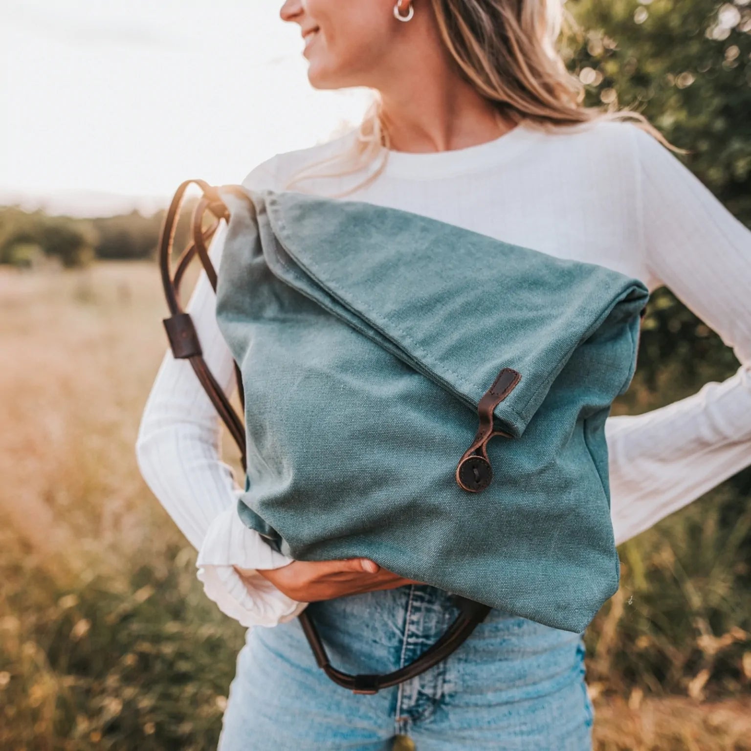 Woman outdoors holding a turquoise canvas handbag with leather strap, wearing jeans and white top.