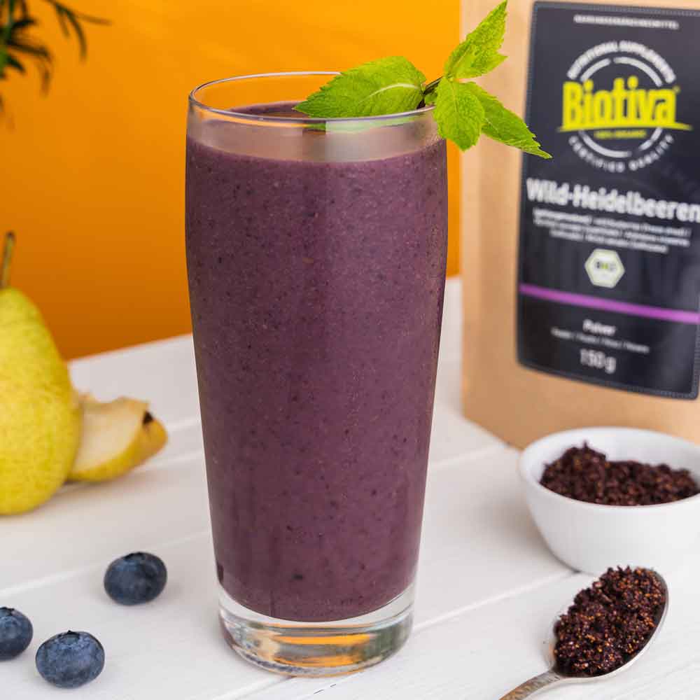 A tall glass of purple smoothie made with Biotiva Wild Blueberry Powder, surrounded by fruit and a spoonful of the powder.