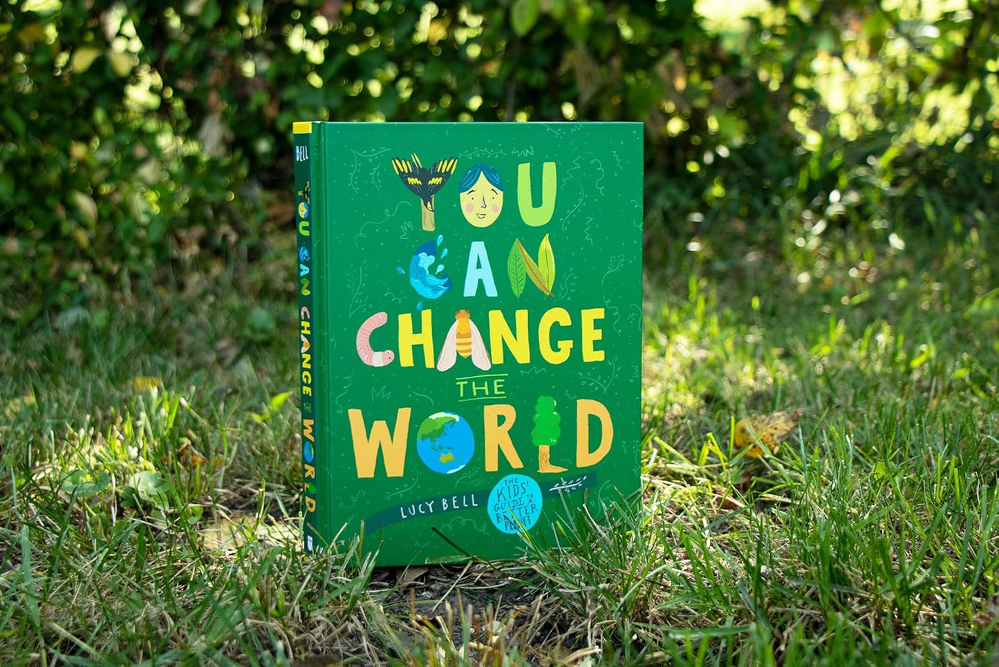 Colorful book titled 'You Can Change the World' on grass with trees in the background