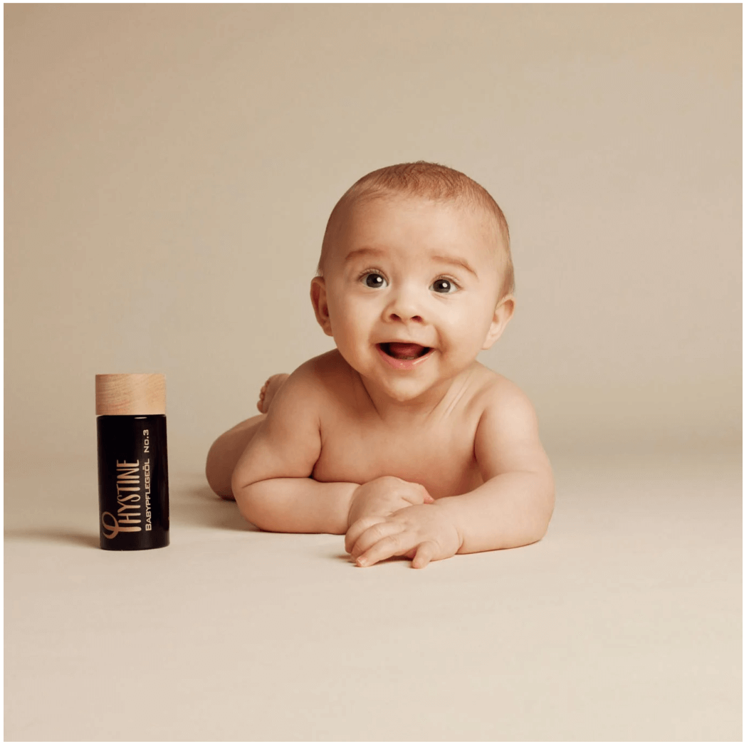 Smiling baby lying on tummy next to Phystine Baby Care Oil No.3 in eco-friendly packaging on neutral background.
