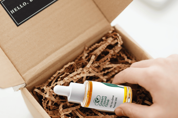Hand holding a bottle of Green People hair oil over an open cardboard box with packing material. For KIND Rituals