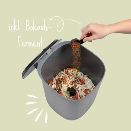 Hand sprinkling Bokashi ferment over food scraps in a grey composting bucket with lid open