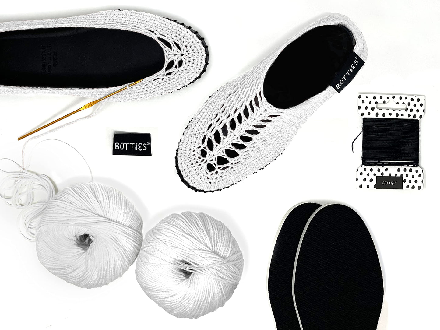 Top view of white crocheted Botties, yarn balls, crochet hook, and black soles ready for crafting.