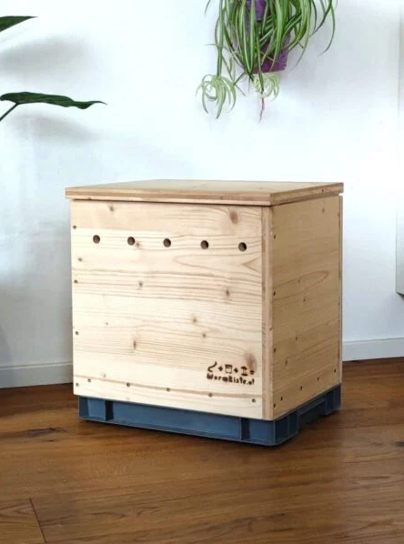 Wooden worm composter box with ventilation holes, placed indoors on a wooden floor, blending with modern home decor.