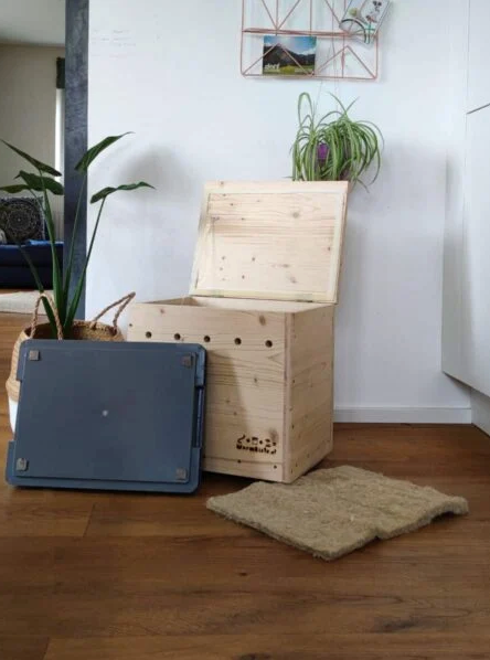 Open wooden worm composting box with lid, tray, and insulating mat displayed on a wooden floor in a cozy indoor setting.