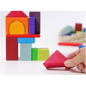 Hand holding a red triangular block, building colorful structures with Grimm’s natural wooden blocks on a white surface.
