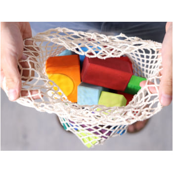 Hands holding a mesh bag filled with colorful Grimm’s natural wooden building blocks seen from above.